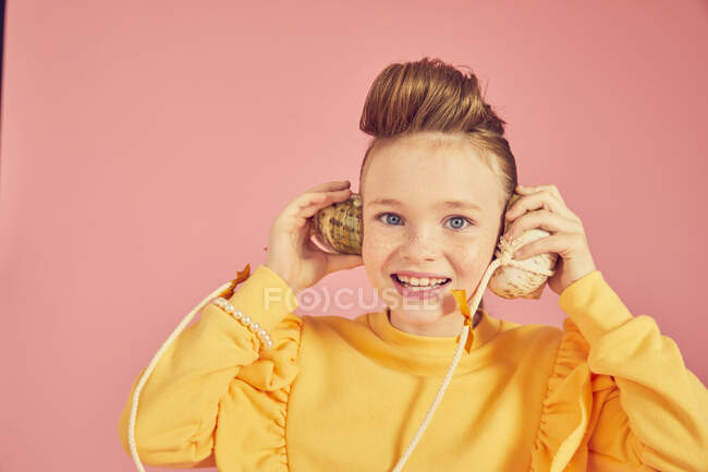 Portrait of brunette girl wearing yellow top, holding sea shell phone, on pink background, looking at camera — Stock Photo