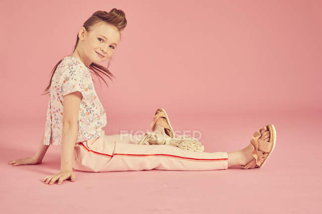 Porträt eines brünetten Mädchens auf rosa Hintergrund, mit floralem Oberteil und blassrosa Hose auf dem Boden sitzend und lächelnd in die Kamera blickend — Stockfoto