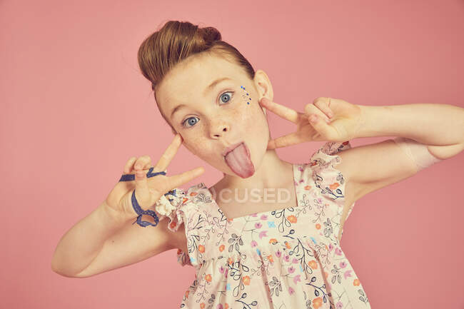 Porträt eines brünetten Mädchens in Rüschenkleid mit Blumenmuster auf rosa Hintergrund, das die Zunge in die Kamera reckt — Stockfoto