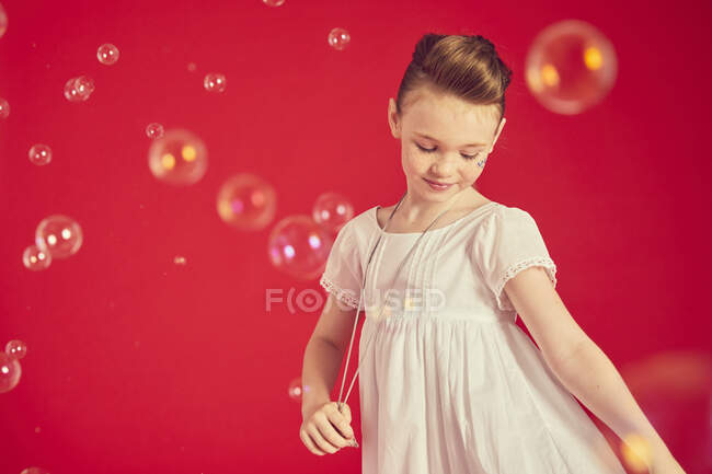 Cute girl wearing romantic white dress on red background, surrounded by soap bubbles — Stock Photo