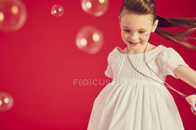 Chica morena vistiendo vestido blanco romántico sobre fondo rojo, rodeado de burbujas de jabón - foto de stock