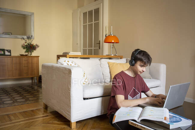 Niño con auriculares sentados en el suelo en la sala de estar, escribiendo en el ordenador portátil, estudiando. - foto de stock
