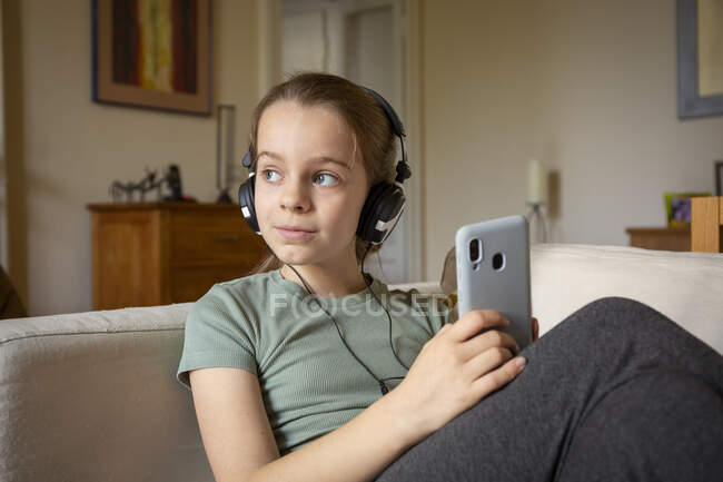 Girl wearing headphones sitting on sofa, holding mobile phone. — Stock Photo