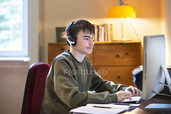 Хлопчик у навушниках сидить за столом перед комп'ютером, вивчаючи . — стокове фото