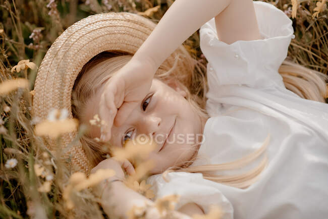 Retrato de menina com cabelo loiro usando chapéu de palha, deitado em um prado. — Fotografia de Stock