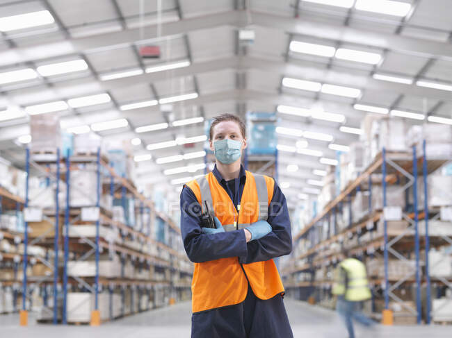 Uomo con maschera chirurgica e gilet ad alta visibilità che lavora in un grande magazzino. — Foto stock