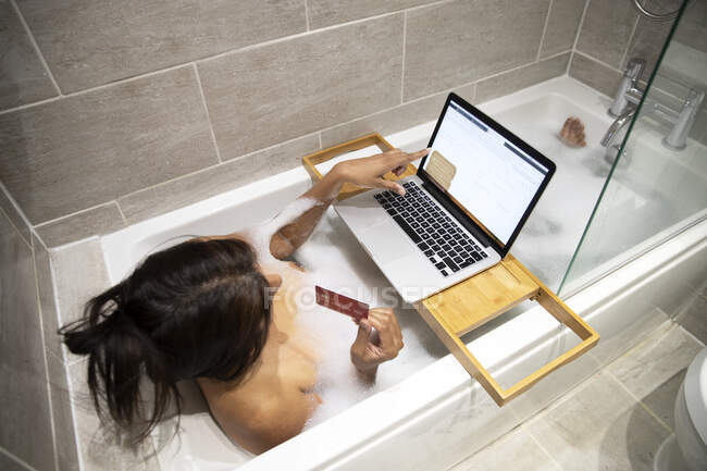 Mujer sentada en la bañera, con baño de espuma y compras en línea en su computadora portátil durante la crisis de Coronavirus. - foto de stock