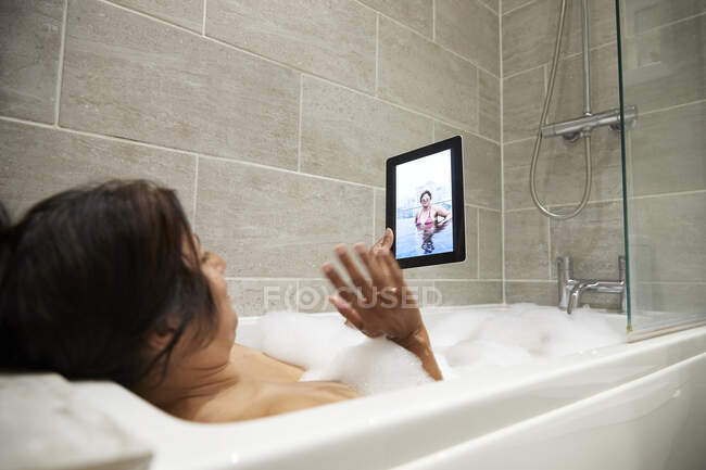 Mujer sentada en la bañera, tomando baño de espuma y usando tableta digital durante la crisis de Coronavirus. - foto de stock