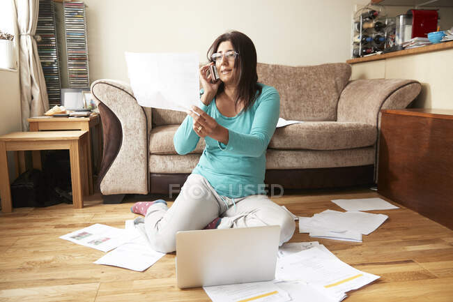 Femme assise sur le sol en bois dans le salon, entourée d'un ordinateur portable et de papiers, travaillant de la maison pendant la crise du coronavirus. — Photo de stock