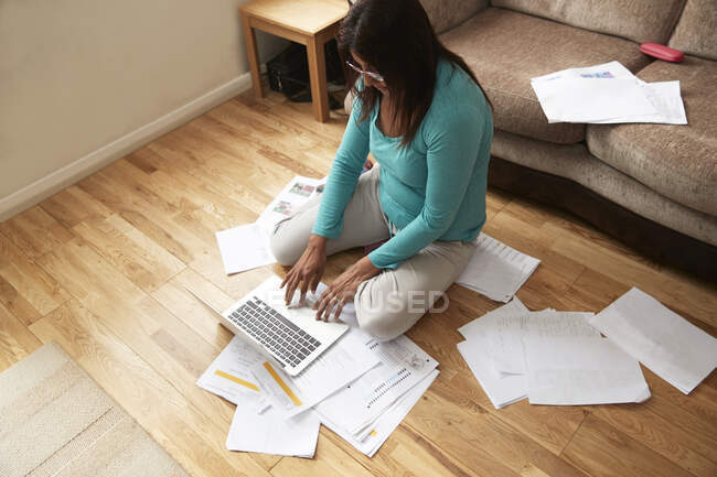 Frau sitzt auf Holzboden im Wohnzimmer, umgeben von Laptop und Papieren, arbeitet während der Coronavirus-Krise von zu Hause aus. — Stockfoto