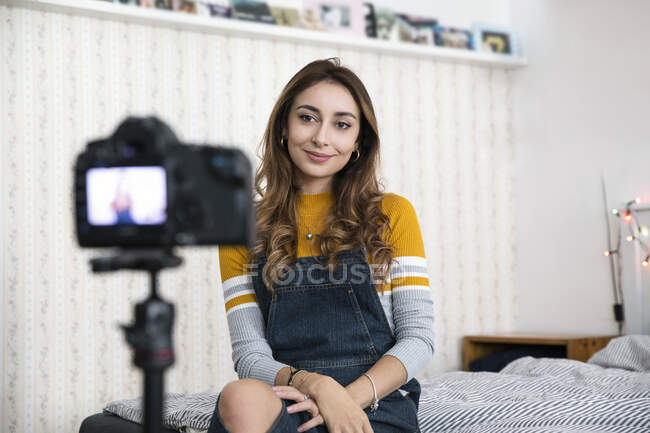 Junge Frau mit langen braunen Haaren sitzt auf dem Bett und nimmt Blog mit Digitalkamera auf. — Stockfoto