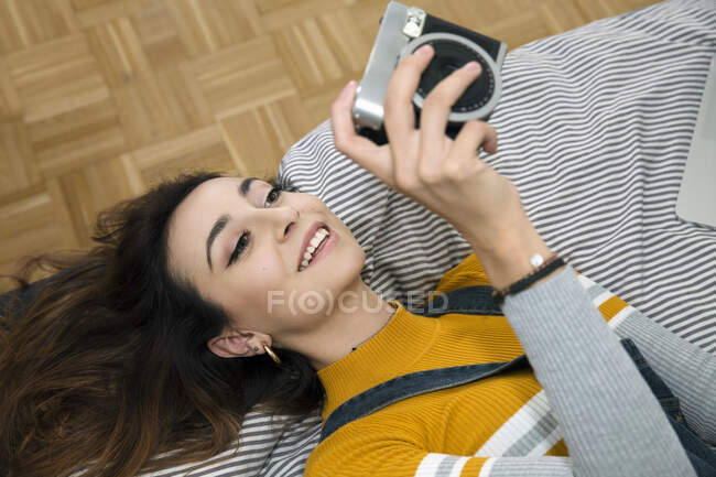 Giovane donna con lunghi capelli castani sdraiata sul letto, scattando selfie con macchina fotografica. — Foto stock