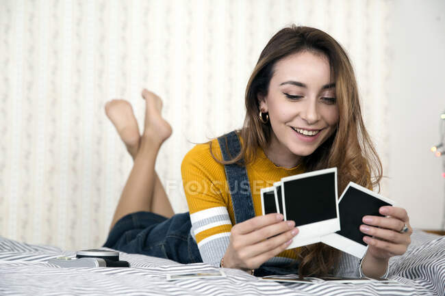 Junge Frau mit langen braunen Haaren liegt auf dem Bett und betrachtet Polaroid-Fotografien. — Stockfoto