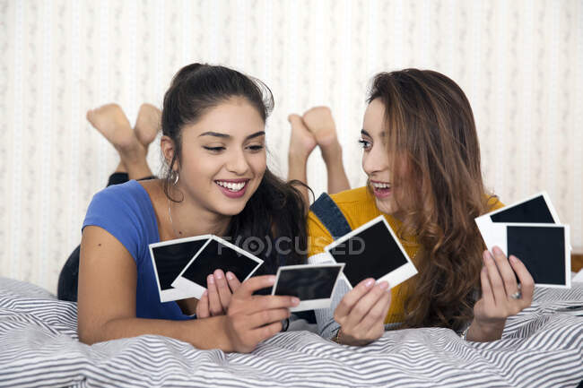 Dos mujeres jóvenes con el pelo castaño largo acostadas en la cama, mirando fotografías Polaroid. - foto de stock