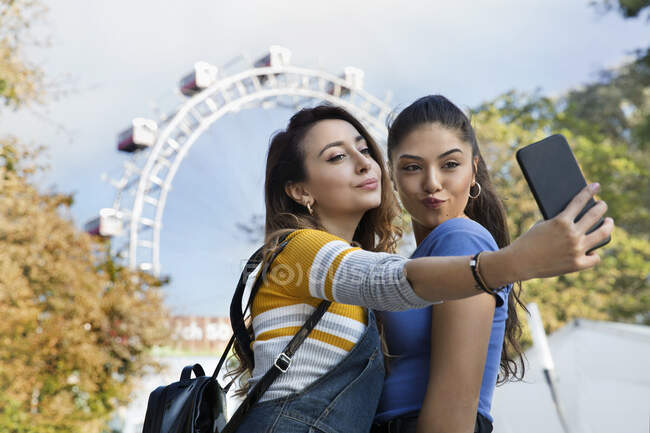 Dos mujeres jóvenes con el pelo castaño largo de pie en un parque cerca de una rueda de la fortuna, tomando selfie con teléfono móvil. - foto de stock