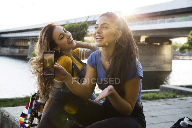 Two young women with long brown hair sitting on riverbank, holding mobile phone and smiling. — Stock Photo
