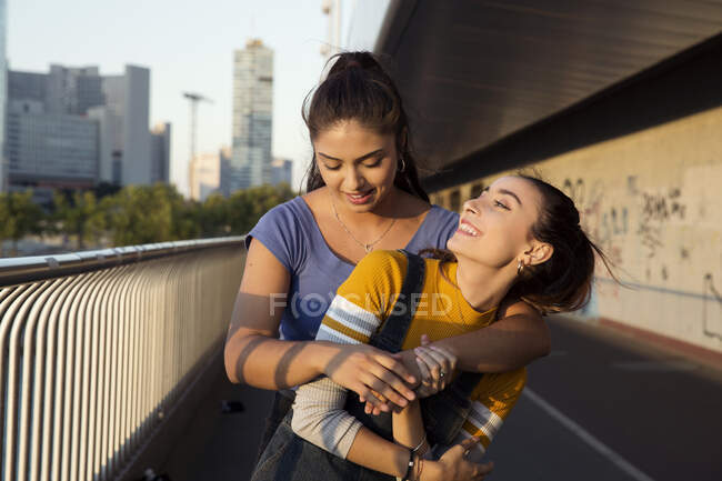 Dos mujeres jóvenes con el pelo castaño largo de pie en el puente urbano, abrazando y sonriendo. - foto de stock