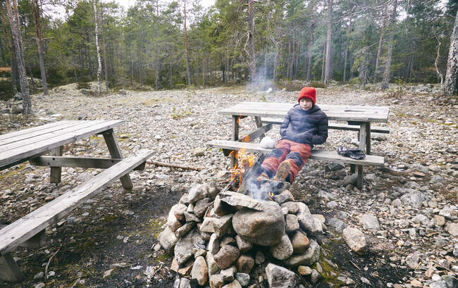 Niño sentado en el banco de picnic junto a la fogata en un bosque en Vasterbottens Lan, Suecia. - foto de stock