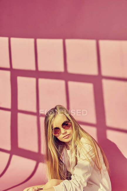 Portrait of girl with long blond hair wearing sunglasses and jacket, on pink background. — Stock Photo