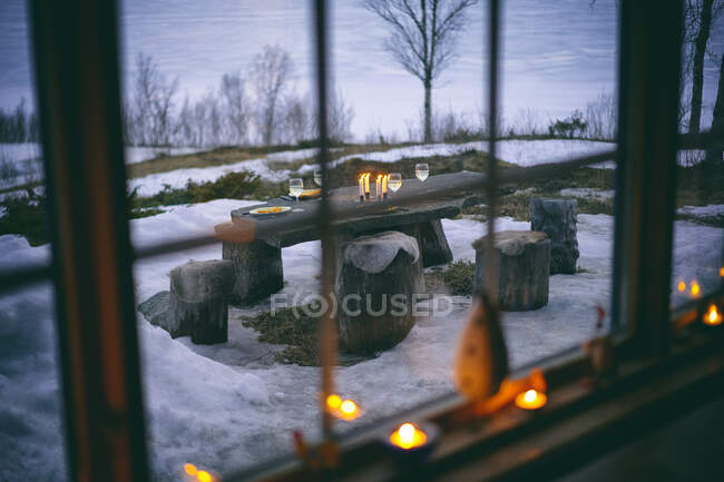 Blick durch das Blockhausfenster auf den Tisch im Freien mit Weingläsern und Kerzen in Vasterbottens Lan, Schweden. — Stockfoto