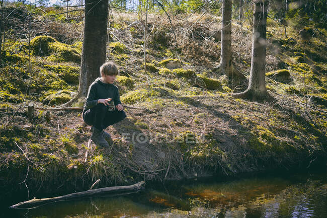 Boy kneeling on riverbank in a forest in Vasterbottens Lan, Sweden. — Stock Photo