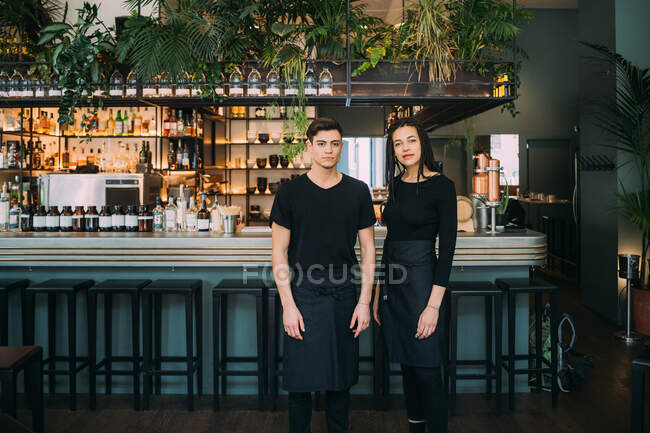Portrait de jeune femme et homme portant des vêtements noirs debout devant le comptoir du bar, souriant à la caméra. — Photo de stock