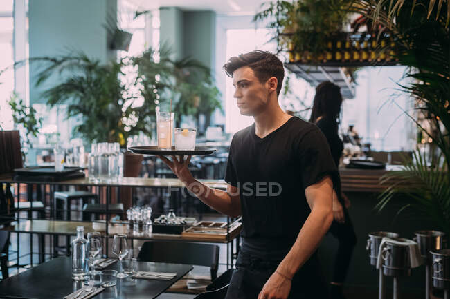 Jovem vestindo roupas pretas trabalhando em bar, carregando bebidas em uma bandeja. — Fotografia de Stock