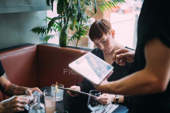 Camarero de pie en una mesa en un bar, tomando el orden usando tableta digital. - foto de stock