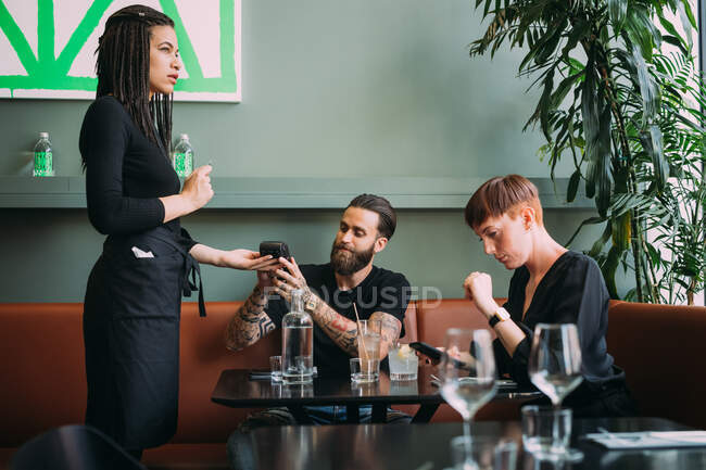 Giovane donna con i capelli corti e barbuto giovane seduto in un bar, utilizzando il telefono cellulare per pagare. — Foto stock