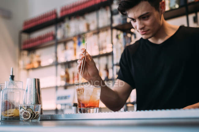 Young man wearing black clothes standing behind bar counter, preparing drink. — Stock Photo
