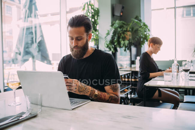 Young bearded man wearing black T-Shirt sitting at table in a bar, using mobile phone and laptop computer. — Stock Photo