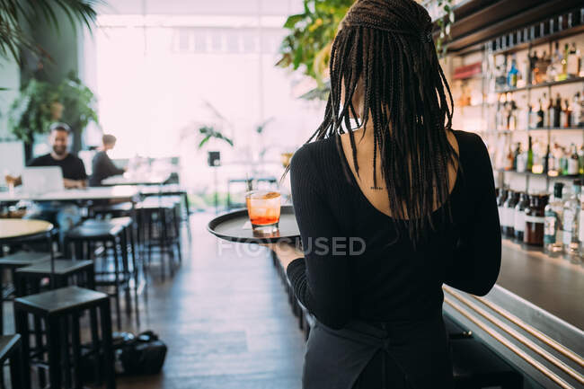 Vue arrière de serveuse vêtue de vêtements noirs travaillant dans un bar, transportant des boissons sur un plateau. — Photo de stock