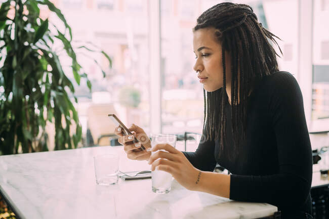 Mujer joven con rastas, vistiendo camiseta negra sentada a la mesa en un bar, usando teléfono móvil. - foto de stock