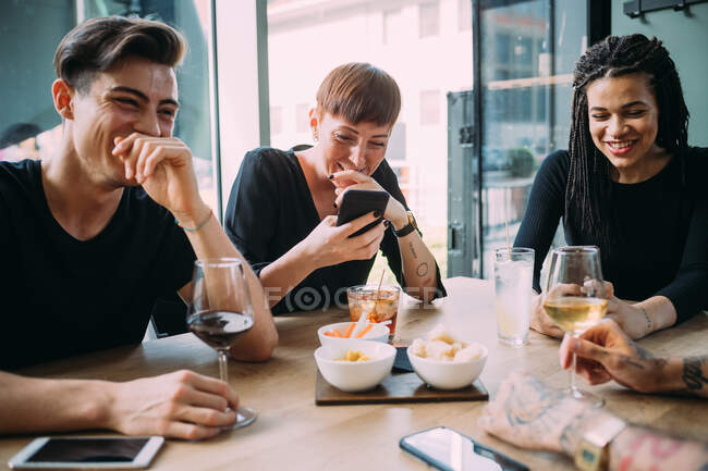 Deux jeunes femmes et hommes vêtus de vêtements décontractés assis à une table dans un bar, riant. — Photo de stock
