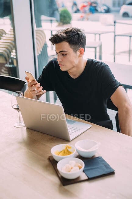 Jovem vestindo camiseta preta sentada à mesa em um bar, usando telefone celular e computador portátil. — Fotografia de Stock