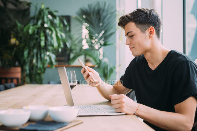 Young man wearing black T-Shirt sitting at table in a bar, using mobile phone and laptop computer. — Stock Photo
