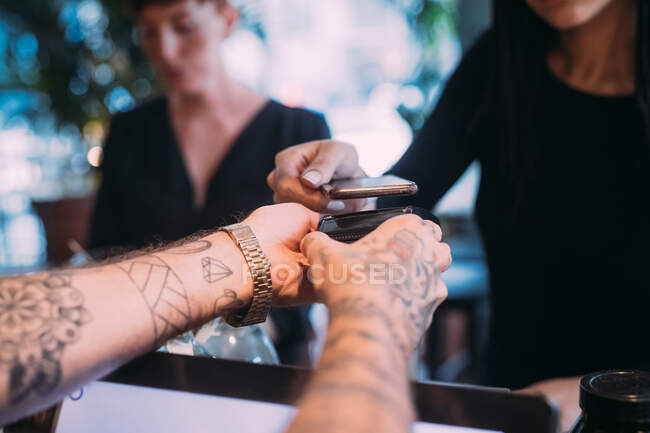 Gros plan du barman tatoué debout au comptoir, distribuant un lecteur de carte à une femme utilisant un téléphone portable pour payer. — Photo de stock