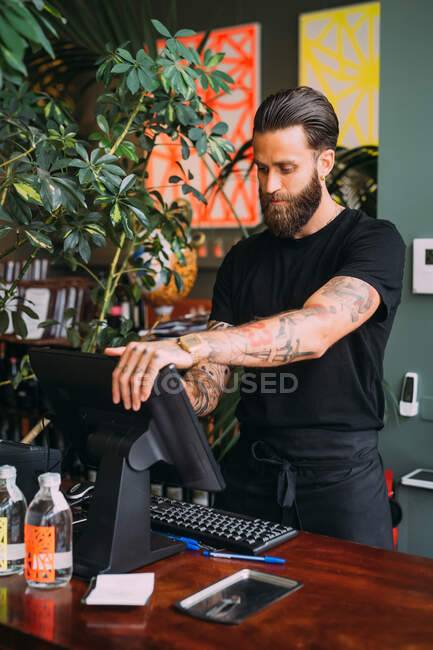 Bearded, tattooed bartender standing at counter, using computerized till. — Stock Photo