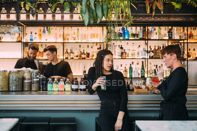 Portrait of two young women sitting at a bar counter and two young men, working behind bar. — Stock Photo
