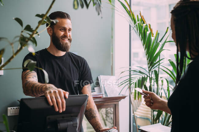 Bearded, tattooed bartender standing at counter, smiling at woman holding credit card. — Stock Photo