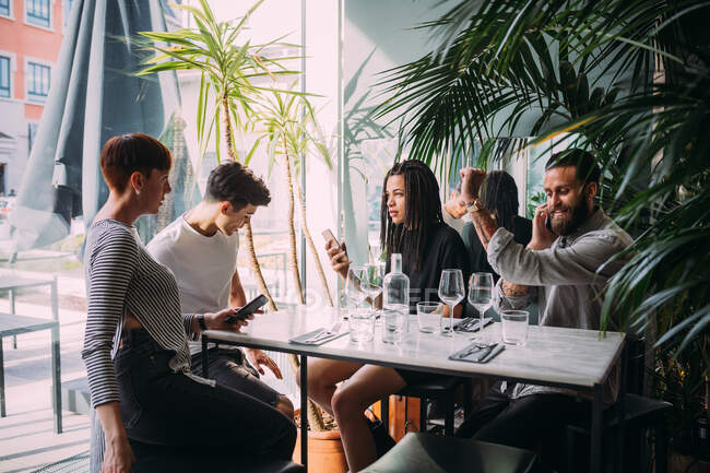 Дві молоді жінки і чоловіки в повсякденному одязі сидять за столом в барі, використовуючи свої мобільні телефони . — стокове фото