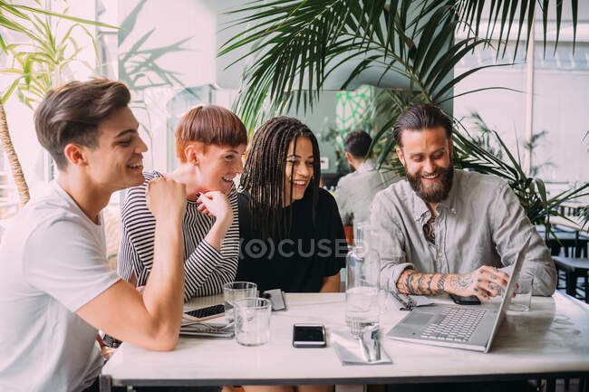 Zwei junge Frauen und Männer in lässiger Kleidung sitzen an einem Tisch in einer Bar und schauen auf Laptop. — Stockfoto