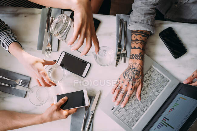 Высокоугольный крупный план четырех человек, сидящих за столом в баре, с помощью мобильных телефонов и ноутбука. — стоковое фото