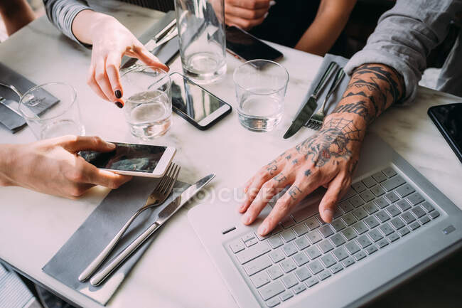 Gran angular primer plano de cuatro personas sentadas en una mesa en un bar, utilizando teléfonos móviles y portátil. - foto de stock