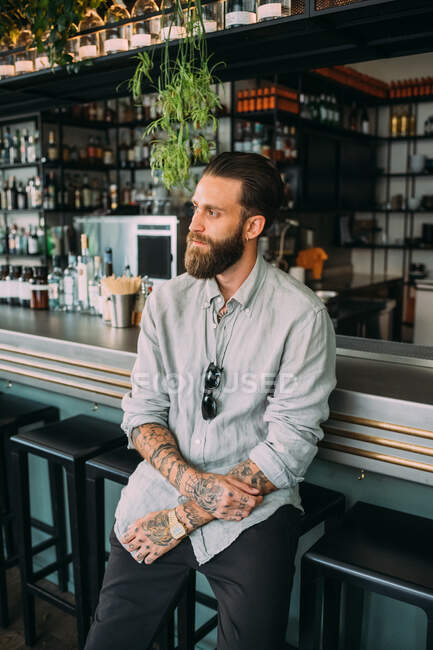 Portrait of bearded young man with brown hair, with tattoos on arms, wearing grey shirt, sitting at a bar counter. — Stock Photo