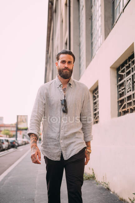 Portrait de jeune homme barbu aux cheveux bruns, tatoué sur les bras, portant une chemise grise, marchant dans la rue. — Photo de stock