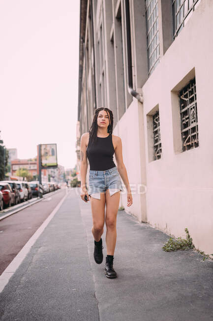 Ritratto di giovane donna dai capelli castani scuri, vestita con gilet nero e pantaloncini di jeans, che cammina per strada. — Foto stock