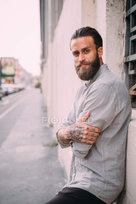 Portrait of bearded young man with brown hair, with tattoos on arms, wearing grey shirt, leaning against wall. — Stock Photo