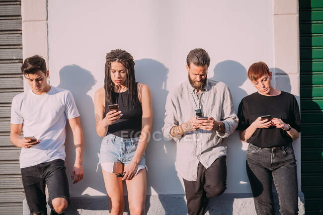 Дві молоді жінки і чоловіки в повсякденному одязі, спираючись на стіну, використовуючи мобільні телефони . — стокове фото