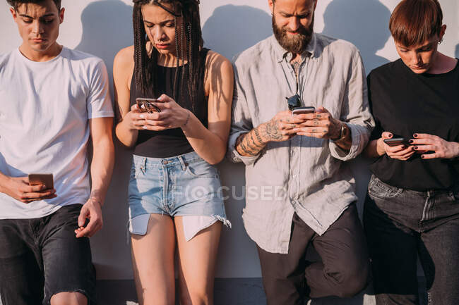 Due giovani donne e uomini che indossano abiti casual appoggiati al muro, utilizzando i telefoni cellulari. — Foto stock